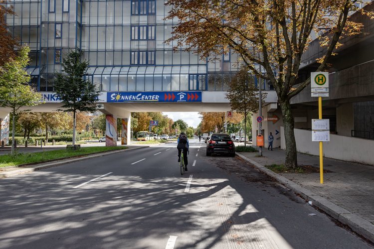 Breite Straße mit parkenden Autos, Bäumen, einer Bushaltestelle und einer Radfah