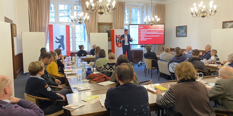 Viele Menschen sitzen an Tischen und hören einer Person zu