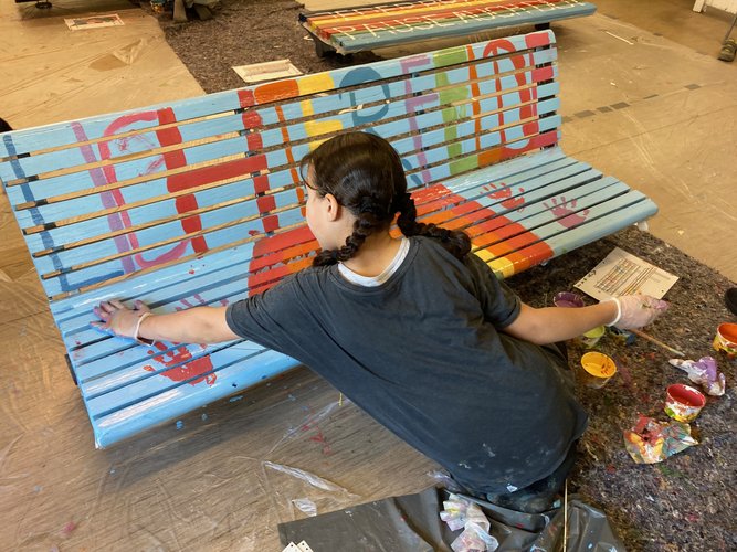 Ein Kind bemalt mit Farben eine Bank.