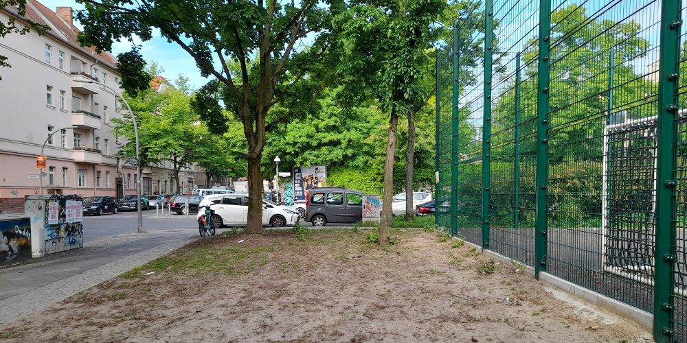 sandiger Platz neben einem Sportplatz, mit Bäumen und Wohnhäusern im Hintergrund