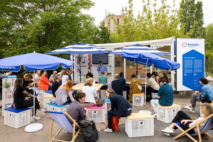 Ein umgebauter, offener Container und davor Menschen die einem Vortrag zuhören.