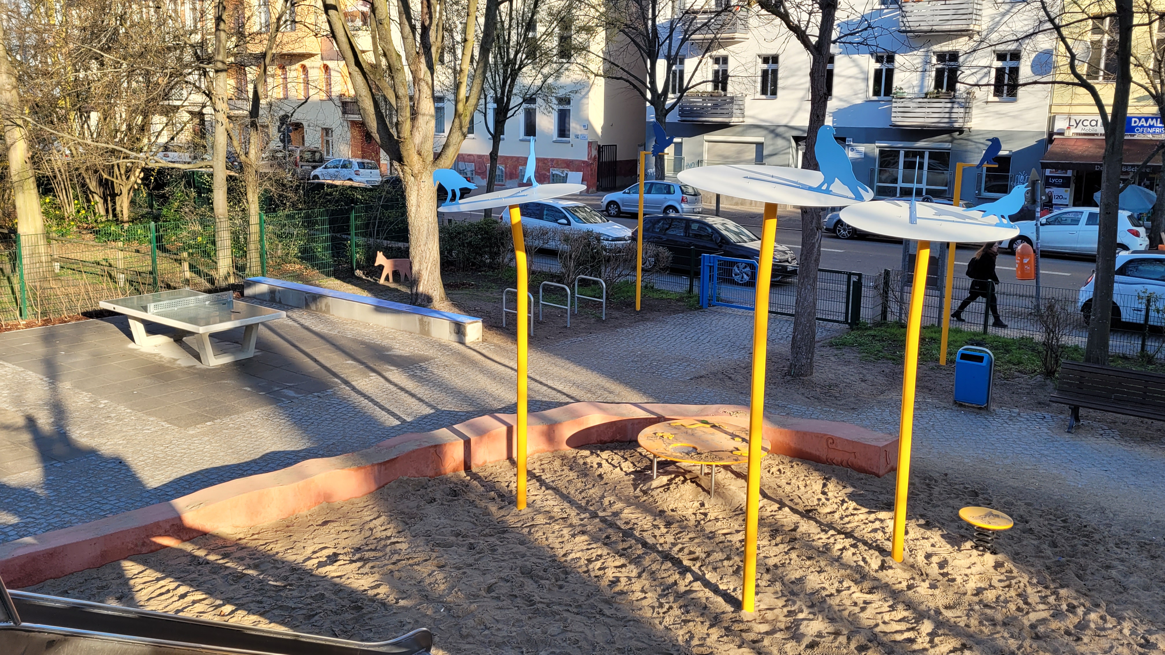 Spielplatz, Sandkasten, Rutsche und eine Tischtennisplatte. Bäume, Straße und Wohnhäuser im Hintergrund