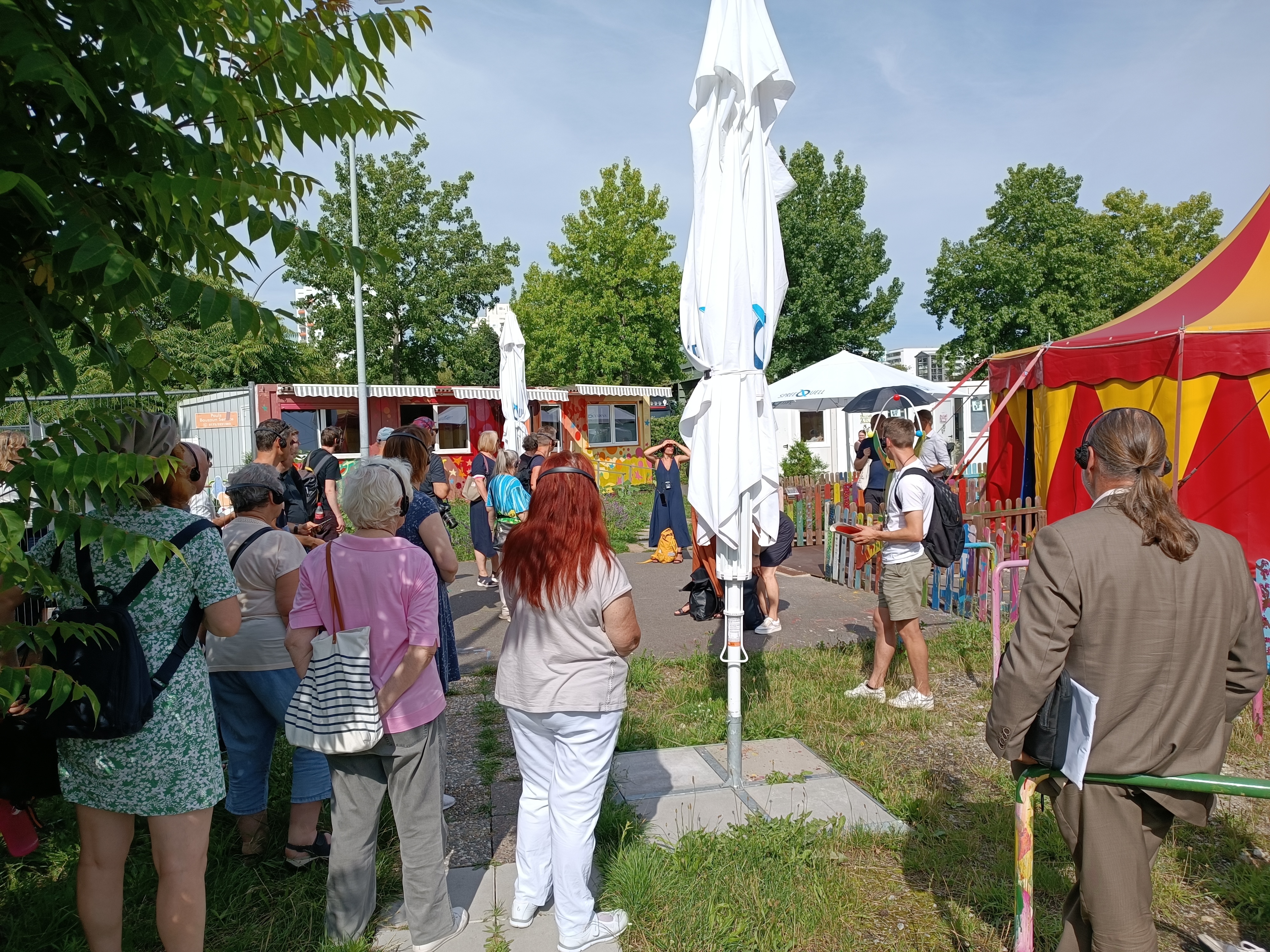 Gruppe von Personen auf einem Platz vor dem Zirkus Cabuwazi mit Kopfhörern, die den Sprechern des Rundgangs zuhören